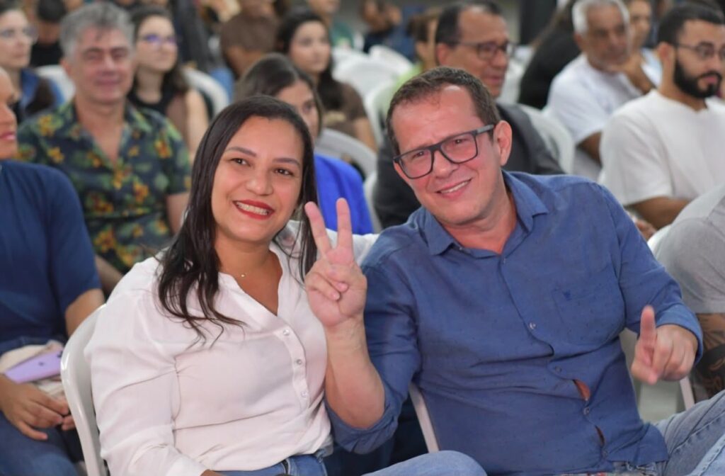 Fabinho Taciano sentado na plateia da convenção, ao lado de uma mulher; os dois fazem sinal de V com os dedos